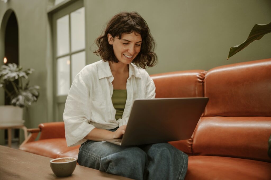 A young woman with pale skin and short, cropped brown hair sits in front of her laptop smiling, seated on a rust leather sofa in a room with painted sage green walls.