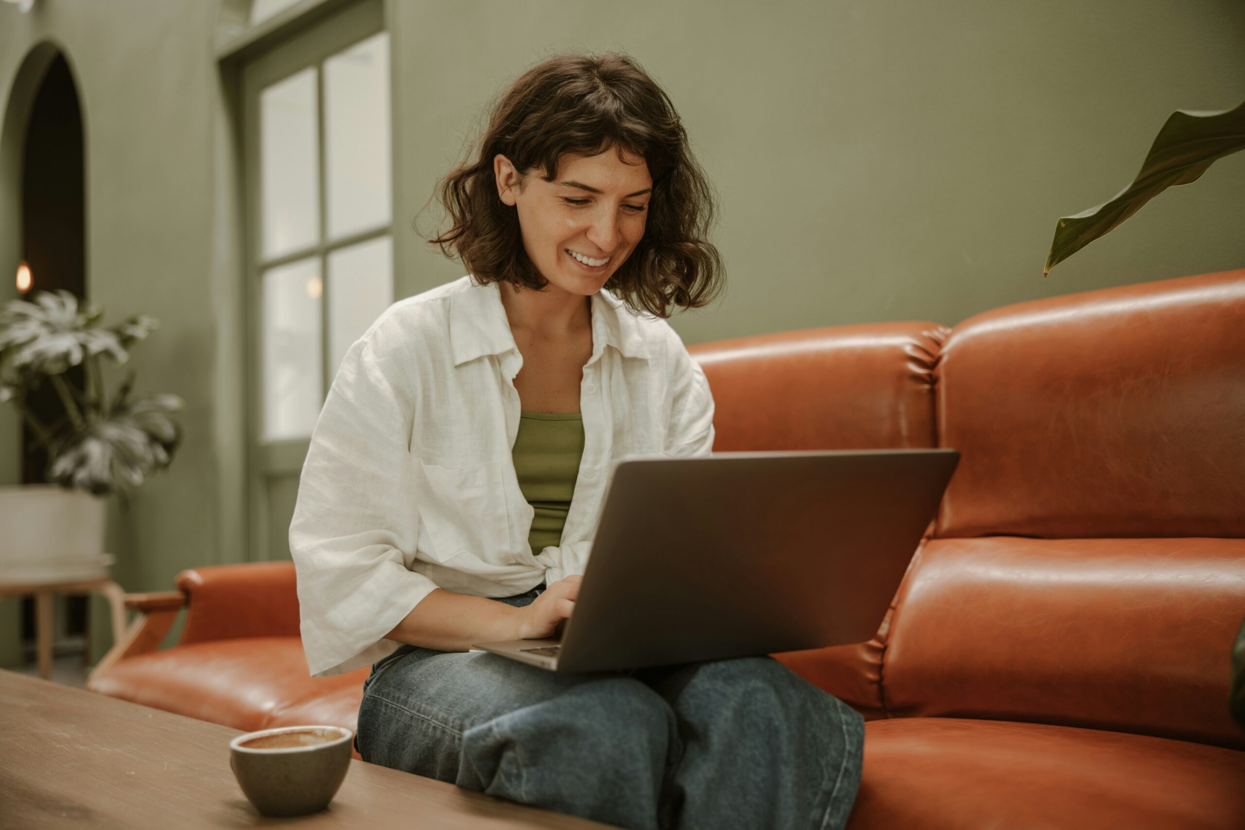 A young woman with pale skin and short, cropped brown hair sits in front of her laptop smiling, seated on a rust leather sofa in a room with painted sage green walls.